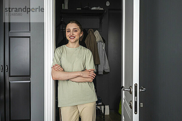 Lächelnde junge Frau steht mit verschränkten Armen vor der Tür