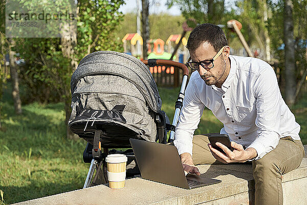 Männlicher Unternehmer  der ein Mobiltelefon hält  während er einen Laptop an einem Kinderwagen im Park benutzt