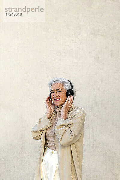 Nachdenkliche reife Frau lächelt  während sie über Kopfhörer vor einer Wand Musik hört