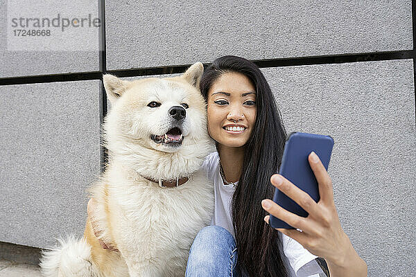 Hipster-Frau macht Selfie mit Akita-Hund durch Handy an der Wand