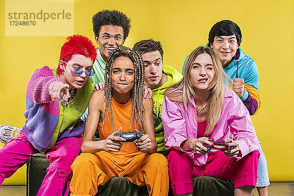 Frauen halten Gamepad  während sie mit Freunden vor einem gelben Hintergrund sitzen