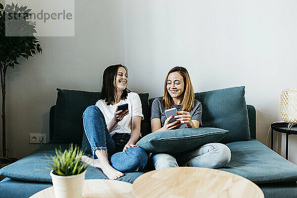 Lächelnde Freundinnen  die zu Hause auf dem Sofa sitzen und ein Mobiltelefon benutzen