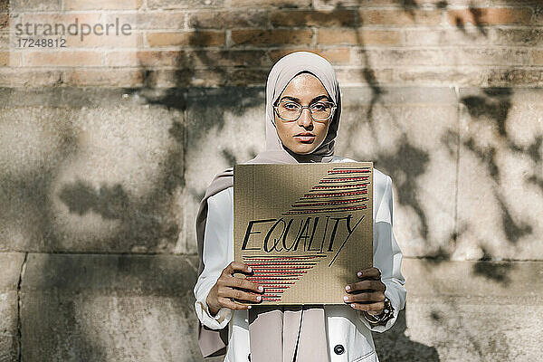 Junge Frau hält Plakat mit Gleichstellungstext vor einer Mauer an einem sonnigen Tag