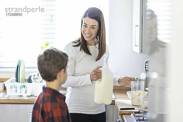 Lächelnde Frau im Gespräch mit ihrem Sohn bei der Zubereitung eines Milchshakes in der Küche