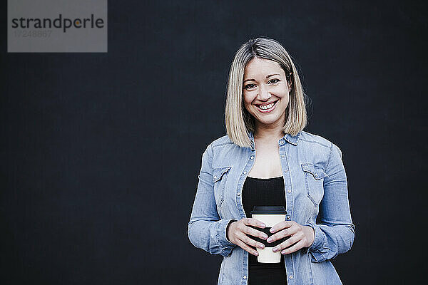 Geschäftsfrau lächelt  während sie eine Einweg-Kaffeetasse vor einer schwarzen Wand hält