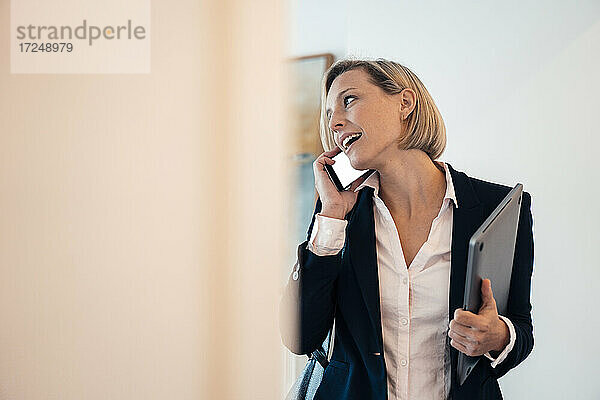 Lächelnde Geschäftsfrau  die einen Laptop hält  während sie zu Hause telefoniert