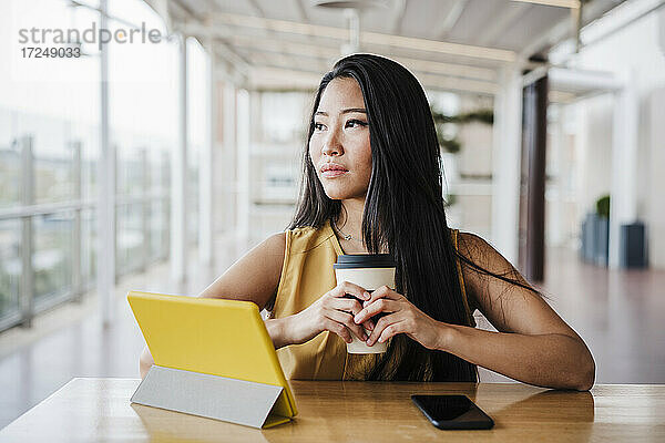 Geschäftsfrau schaut weg  während sie eine Kaffeetasse in einer modernen Büro-Cafeteria hält