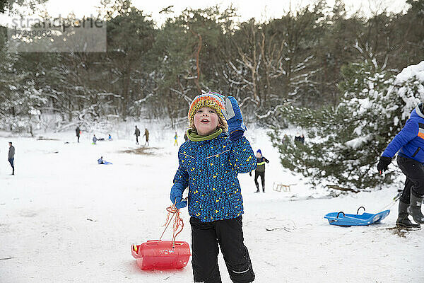 Verspielter Junge mit Schlitten auf Schnee stehend