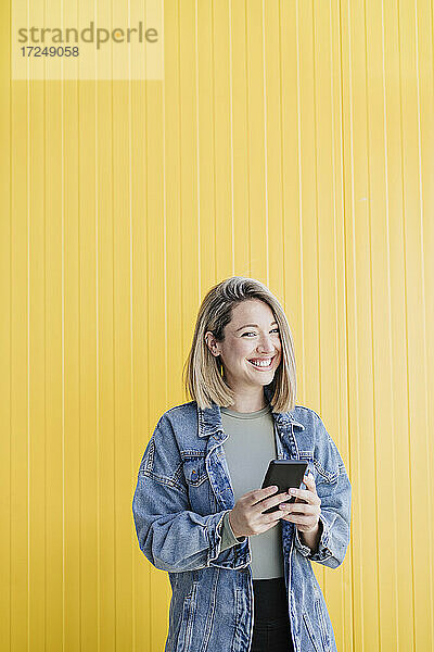Lächelnde Frau mit Mobiltelefon vor einer Wand stehend