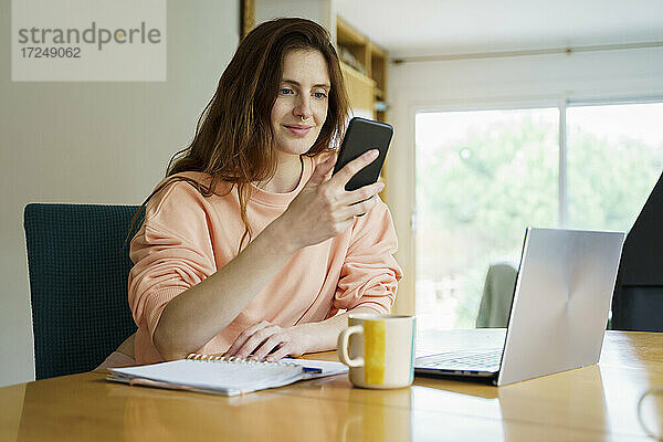 Frau benutzt Smartphone  während sie mit Laptop und Kaffeetasse am Tisch sitzt