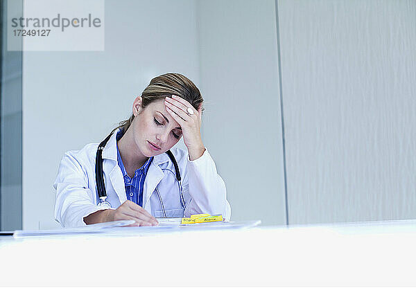 Gestresste Ärztin mit Kopf in der Hand am Schreibtisch sitzend im Krankenhaus