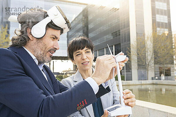 Männlicher Ingenieur mit Virtual-Reality-Headset  der das Modell einer Windkraftanlage einstellt  während eine Kollegin daneben steht