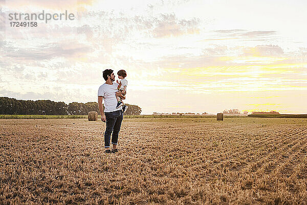 Vater trägt seinen kleinen Sohn durch ein abgeerntetes Feld bei Sonnenuntergang