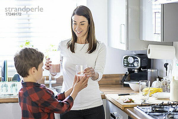 Lächelnde Mutter gibt ihrem Sohn in der Küche zu Hause ein Glas Milchshake