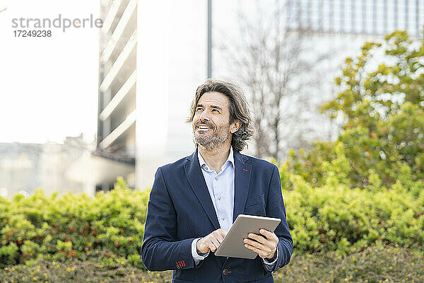 Männlicher Fachmann mit digitalem Tablet  der wegschaut  während er im Büropark steht