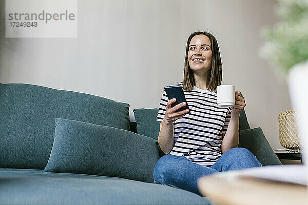 Lächelnde Frau mit Kaffee und Handy in der Hand auf dem Sofa im Ferienhaus
