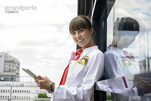 Lächelnde Ärztin mit digitalem Tablet  die sich an eine Glaswand auf einem Balkon lehnt