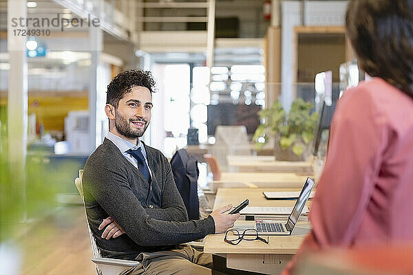 Lächelnder Geschäftsmann mit Mobiltelefon  der eine Kollegin im Büro anschaut
