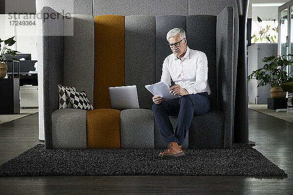 Männlicher Unternehmer liest ein Dokument  während er im Büro am Laptop sitzt