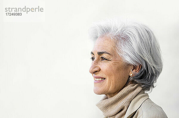 Lächelnde Frau mit grauem Haar  die an einer weißen Wand nachdenkt