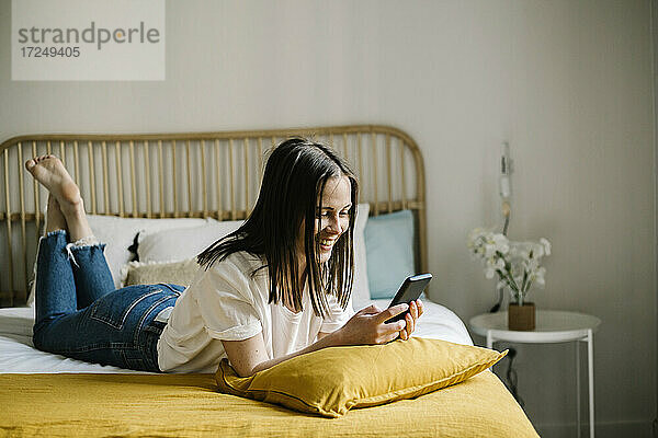 Lächelnde Frau  die ein Smartphone benutzt  während sie zu Hause auf dem Bett liegt