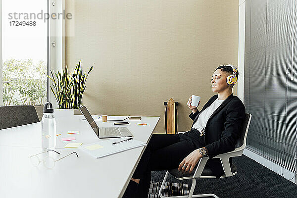 Geschäftsfrau mit Kopfhörern und Kaffeetasse vor einem Laptop im Büro