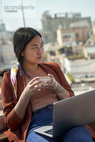 Nachdenkliche Frau sitzt mit Laptop und Kaffeetasse auf der Dachterrasse