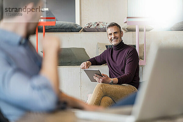 Lächelnder Geschäftsmann schaut auf einen männlichen Kollegen  der ein digitales Tablet im Büro hält