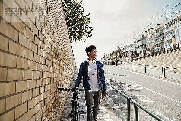 Lächelnder gut aussehender Mann schaut weg  während er mit dem Fahrrad auf dem Gehweg spazieren geht