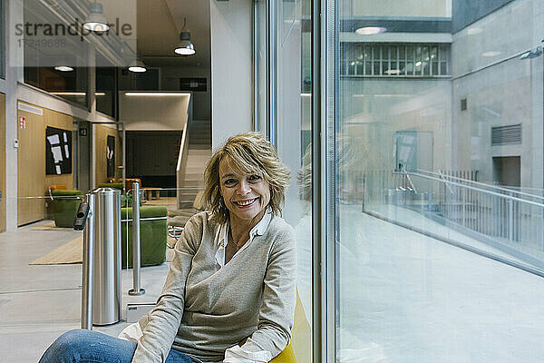 Lächelnde Geschäftsfrau sitzt am Fenster in einem Coworking-Büro