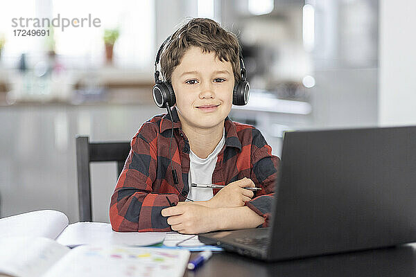 Lächelnder Junge mit Kopfhörern  der zu Hause vor einem Laptop sitzt