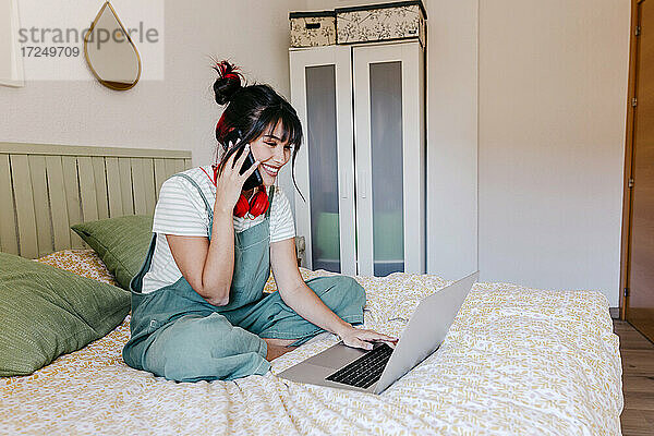 Junge Frau spricht mit dem Handy  während sie einen Laptop im Schlafzimmer benutzt