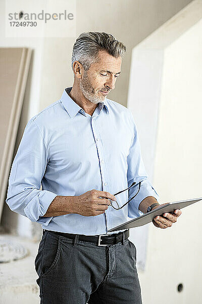 Männlicher Architekt  der eine Brille hält und auf ein digitales Tablet auf einer Baustelle schaut