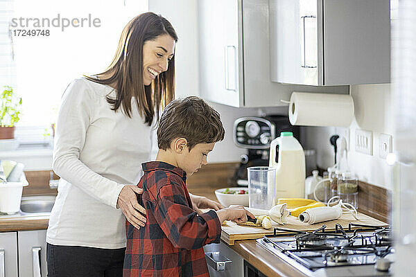 Lächelnde Mutter hilft ihrem Sohn beim Schneiden von Bananen in der Küche zu Hause