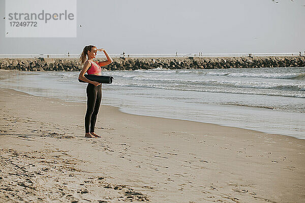 Junge Frau  die ihre Augen abschirmt  während sie eine Übungsmatte am Strand hält