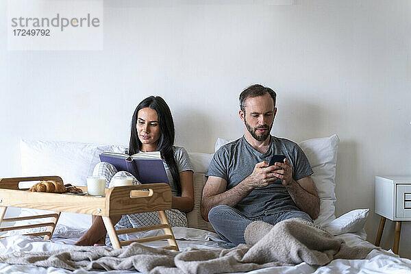 Frau  die ein Buch liest und mit einem Mann zusammensitzt  der ein Smartphone benutzt  während sie zu Hause auf dem Bett frühstückt