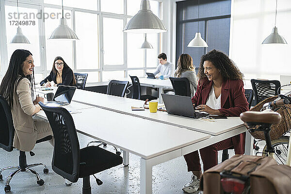Lächelnde Unternehmerinnen  die am Schreibtisch in einem Coworking-Büro diskutieren