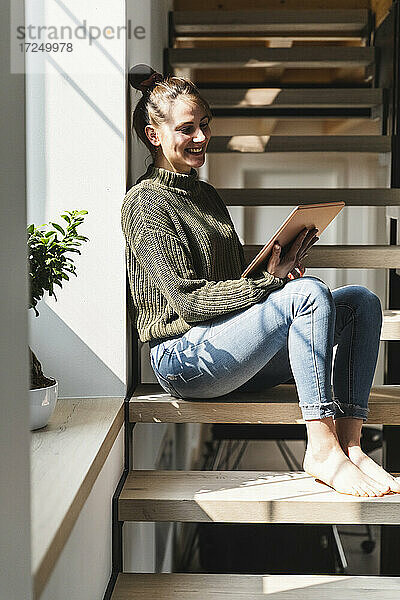 Lächelnde Frau  die ein Tablet benutzt  während sie auf einer Treppe zu Hause sitzt