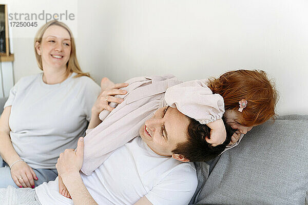 Lächelnde schwangere Frau  die ihre Tochter betrachtet  die mit ihrem Vater auf dem Sofa zu Hause spielt