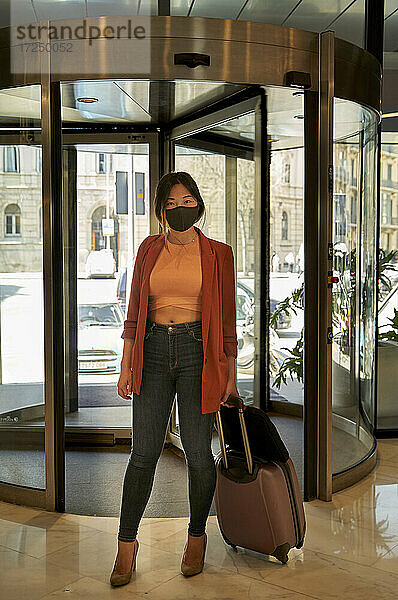 Frau mit Gesichtsschutzmaske steht mit Koffer am Hoteleingang