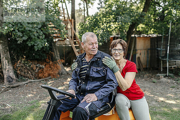 Lächelndes älteres Paar sitzt auf einem Rasenmäher im Hinterhof
