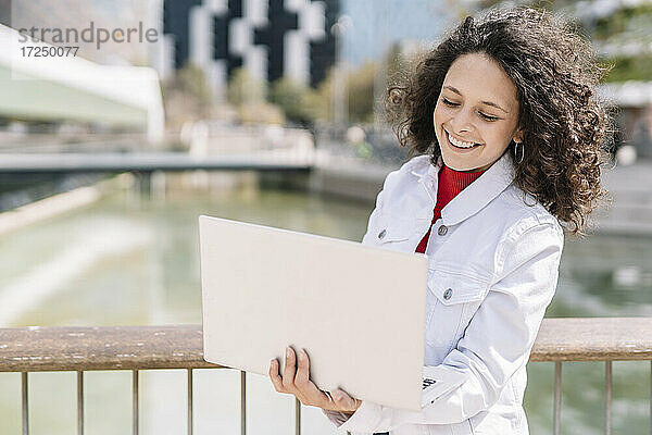 Lächelnde Frau mit Laptop am Geländer an einem sonnigen Tag