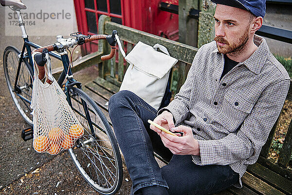 Nachdenklicher Mann mit Mobiltelefon auf einer Bank am Fahrrad sitzend