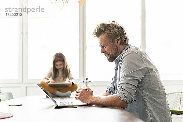 Reifer Geschäftsmann arbeitet am Laptop  während das Mädchen im Hintergrund zu Hause sitzt