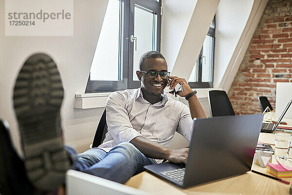 Männlicher Unternehmer  der mit seinem Smartphone spricht  während er einen Laptop im Büro benutzt