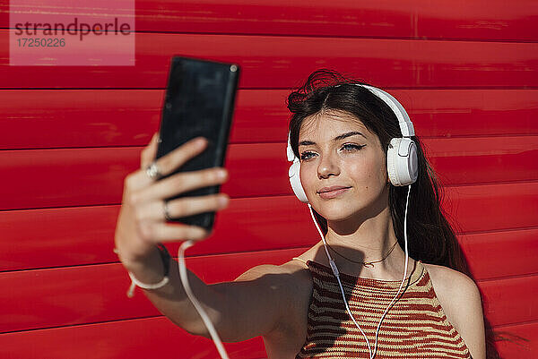 Lächelndes Teenager-Mädchen  das ein Selfie über ein Smartphone macht  während es Musik hört