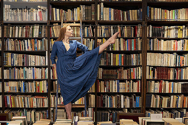 Junge Frau in blauem Kleid balanciert auf einem Bein auf einer Leiter in einer Bibliothek