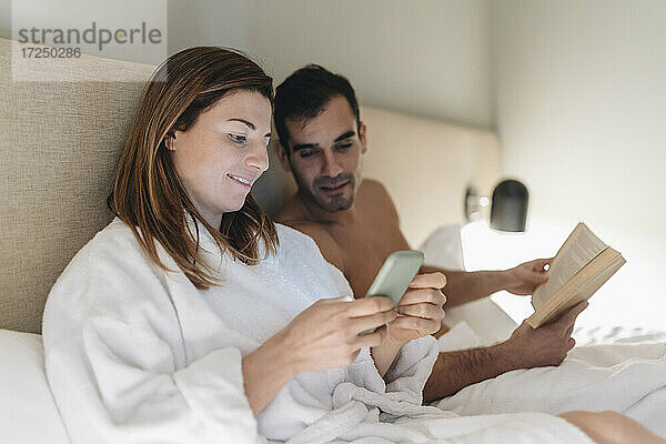 Schöne Frau  die telefoniert  während sie neben einem Mann mit einem Buch auf dem Bett sitzt