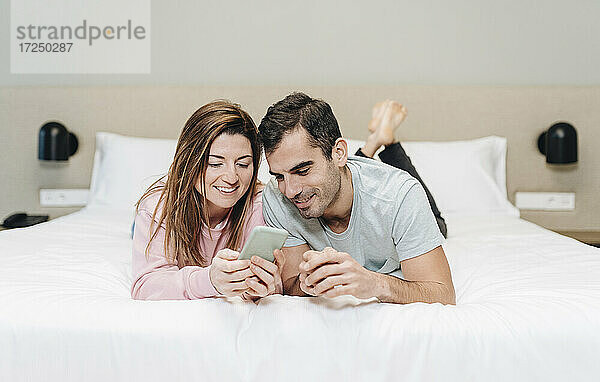 Mid adult couple using mobile phone while lying on bed in hotel