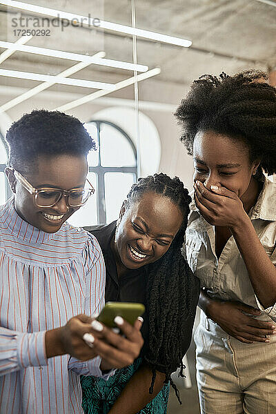 Glückliche weibliche Fachkraft  die ein Mobiltelefon benutzt  während ihre Kolleginnen im Coworking-Büro lachen